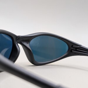 Pode incluir: &Oacute;culos de sol pretos com lentes azuis. Apresentam um design curvo e um acabamento fosco. As hastes s&atilde;o finas e estendem-se da arma&ccedil;&atilde;o. A palavra 'MUTE' &eacute; vis&iacute;vel na arma&ccedil;&atilde;o.