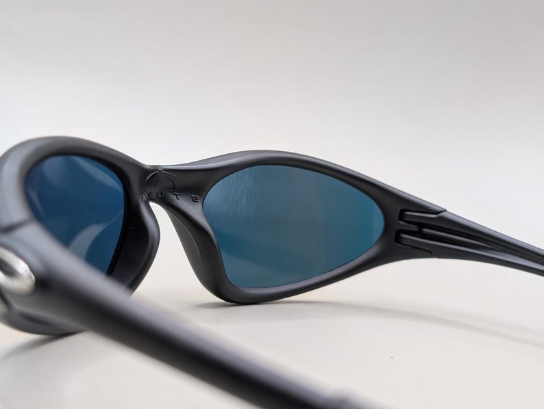 Pode incluir: &Oacute;culos de sol pretos com lentes azuis. Apresentam um design curvo e um acabamento fosco. As hastes s&atilde;o finas e estendem-se da arma&ccedil;&atilde;o. A palavra 'MUTE' &eacute; vis&iacute;vel na arma&ccedil;&atilde;o.
