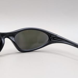May include: Black sunglasses with a curved, streamlined design. The lenses are a dark gray, and the frames are matte black. The temples have a textured detail near the hinges. The sunglasses are designed for sports or outdoor activities.