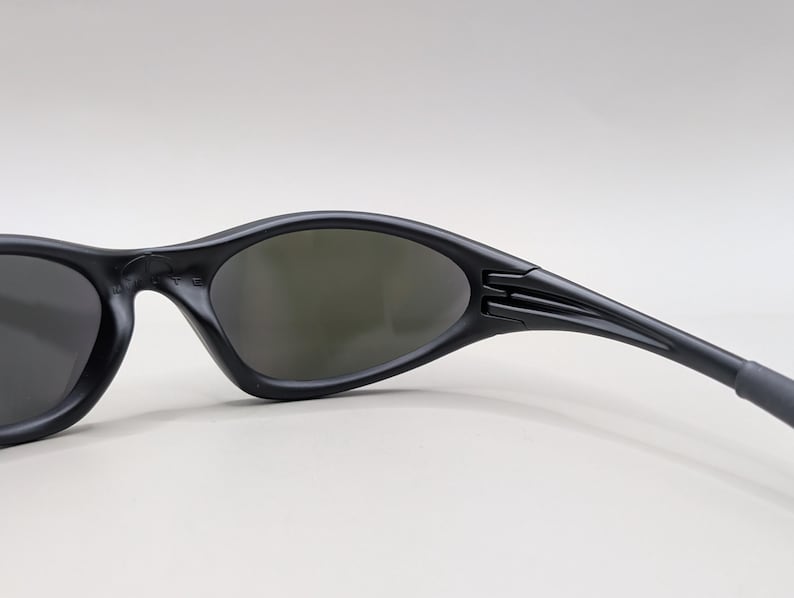 May include: Black sunglasses with a curved, streamlined design. The lenses are a dark gray, and the frames are matte black. The temples have a textured detail near the hinges. The sunglasses are designed for sports or outdoor activities.