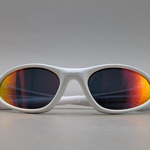 May include: White sunglasses with a curved frame. The lenses feature a gradient effect, shifting from yellow to orange and red. The brand name is visible on the frame. These sunglasses are designed for eye protection and style.