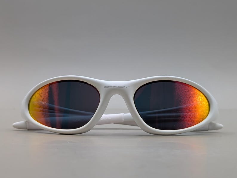 May include: White sunglasses with a curved frame. The lenses feature a gradient effect, shifting from yellow to orange and red. The brand name is visible on the frame. These sunglasses are designed for eye protection and style.