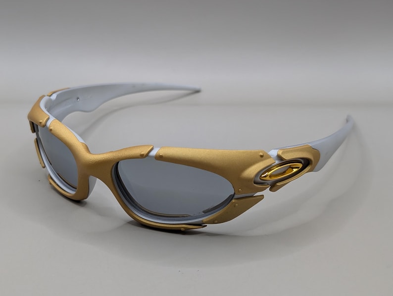 May include: Gold and white sunglasses with gray lenses. The sunglasses have a gold frame around the lenses and gold accents on the temples. The arms are white. The lenses are a dark gray color.
