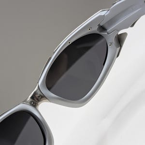 May include: Close-up of silver sunglasses with dark lenses. The sunglasses have a sleek, modern design with a metallic finish. The lenses are a dark gray color, providing shade and protection from the sun.