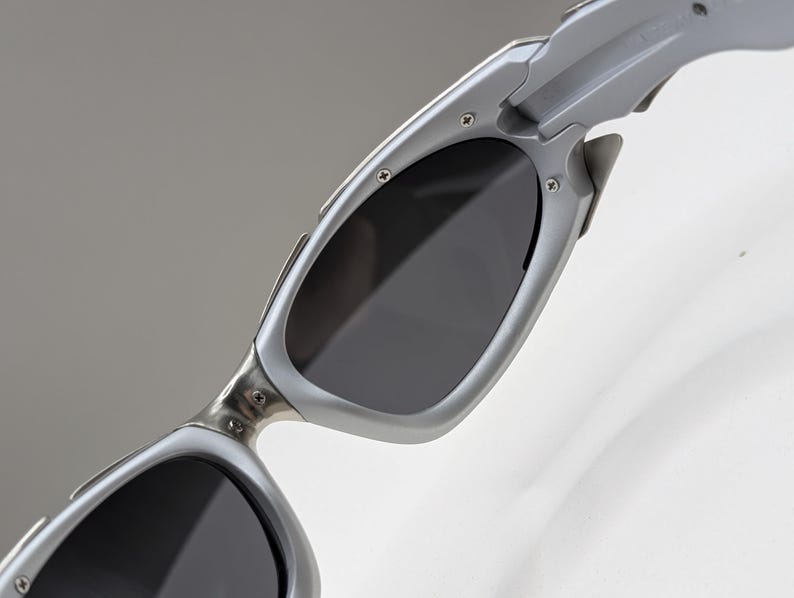 May include: Close-up of silver sunglasses with dark lenses. The sunglasses have a sleek, modern design with a metallic finish. The lenses are a dark gray color, providing shade and protection from the sun.