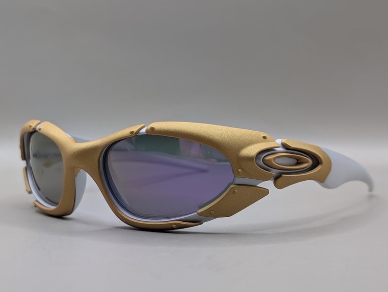 K&ouml;nnte beinhalten: Wei&szlig;e und goldfarbene Sportsonnenbrille mit violett get&ouml;nten Gl&auml;sern. Die Sonnenbrille hat ein futuristisches Design mit einem goldenen Rahmen um die Gl&auml;ser und wei&szlig;en B&uuml;geln. Das Oakley-Logo ist auf dem B&uuml;gel sichtbar.