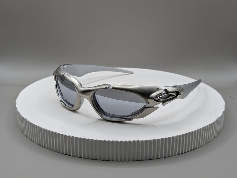 May include: Silver-framed sunglasses with gray lenses. The sunglasses have a futuristic design with a curved frame and a sleek, modern aesthetic. The lenses are a dark gray color, providing a contrast to the silver frame.