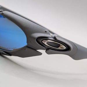 K&ouml;nnte beinhalten: Nahaufnahme einer grauen Sonnenbrille mit blau polarisierten Gl&auml;sern. Die Sonnenbrille hat ein schlankes, modernes Design mit einem silbernen Emblem an der Seite. Die Gl&auml;ser reflektieren einen hellblauen Farbton.