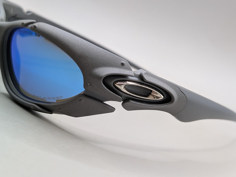 K&ouml;nnte beinhalten: Nahaufnahme einer grauen Sonnenbrille mit blau polarisierten Gl&auml;sern. Die Sonnenbrille hat ein schlankes, modernes Design mit einem silbernen Emblem an der Seite. Die Gl&auml;ser reflektieren einen hellblauen Farbton.