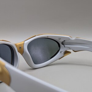 K&ouml;nnte beinhalten: Nahaufnahme einer wei&szlig;en und goldenen Sonnenbrille mit dunklen Gl&auml;sern. Die Sonnenbrille hat ein modernes Design mit einem wei&szlig;en Rahmen und goldenen Akzenten. Die B&uuml;gel sind wei&szlig; und tragen den Schriftzug "MADE IN U.S.A."