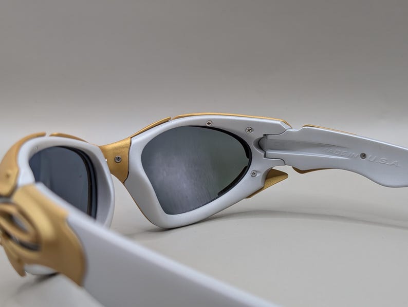 K&ouml;nnte beinhalten: Nahaufnahme einer wei&szlig;en und goldenen Sonnenbrille mit dunklen Gl&auml;sern. Die Sonnenbrille hat ein modernes Design mit einem wei&szlig;en Rahmen und goldenen Akzenten. Die B&uuml;gel sind wei&szlig; und tragen den Schriftzug "MADE IN U.S.A."