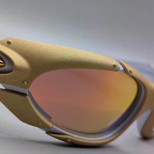 May include: Close-up of white and gold sunglasses with a mirrored lens. The sunglasses have a gold frame with a white accent and a gold hinge. The lens is a gradient of orange and pink.