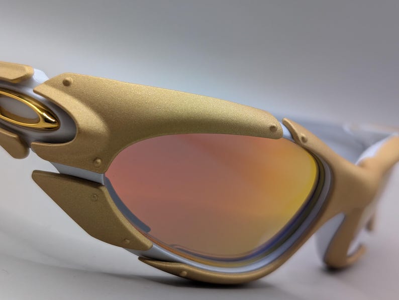 May include: Close-up of white and gold sunglasses with a mirrored lens. The sunglasses have a gold frame with a white accent and a gold hinge. The lens is a gradient of orange and pink.