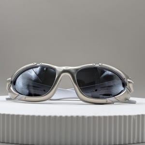 May include: Silver-framed sunglasses with dark lenses. The sunglasses have a futuristic design with a curved frame and a white bridge. The lenses reflect the surrounding environment. The sunglasses are displayed on a white, circular platform.