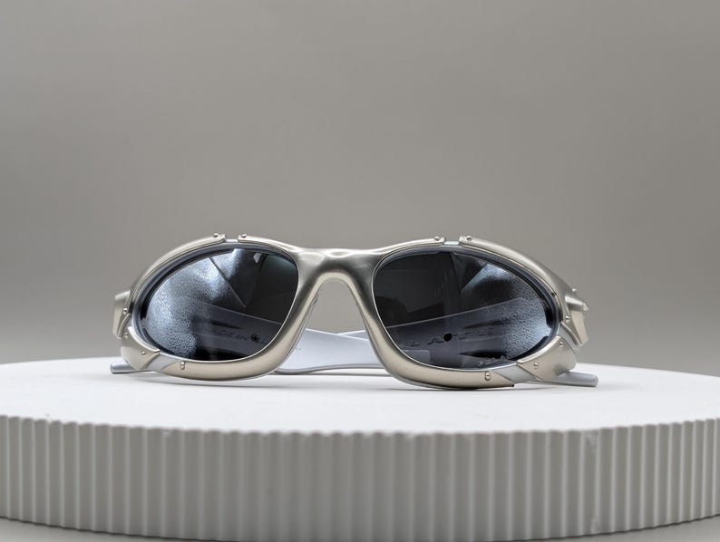 May include: Silver-framed sunglasses with dark lenses. The sunglasses have a futuristic design with a curved frame and a white bridge. The lenses reflect the surrounding environment. The sunglasses are displayed on a white, circular platform.