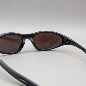K&ouml;nnte beinhalten: Schwarze Sonnenbrille mit braunen Gl&auml;sern. Die Sonnenbrille hat ein schlankes, geschwungenes Design mit matter Oberfl&auml;che. Die B&uuml;gel sind nach hinten gebogen und die Gl&auml;ser sind oval.