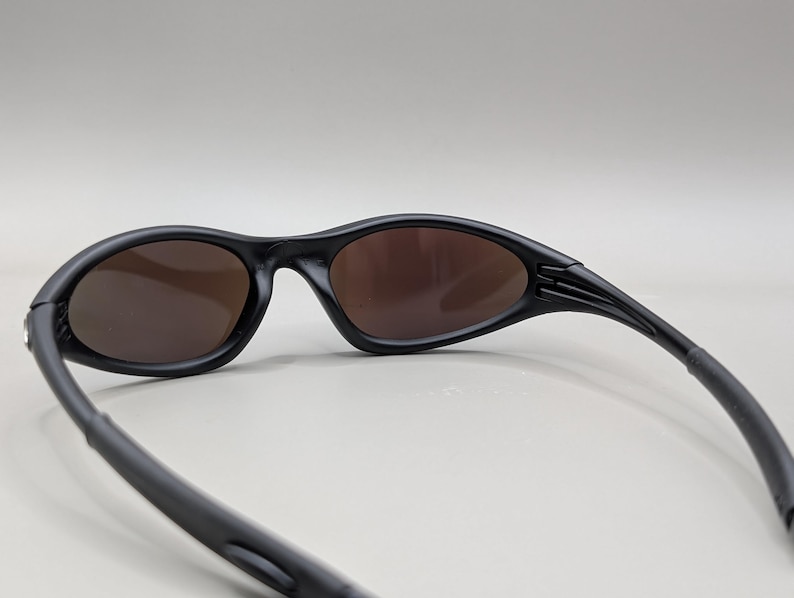 K&ouml;nnte beinhalten: Schwarze Sonnenbrille mit braunen Gl&auml;sern. Die Sonnenbrille hat ein schlankes, geschwungenes Design mit matter Oberfl&auml;che. Die B&uuml;gel sind nach hinten gebogen und die Gl&auml;ser sind oval.