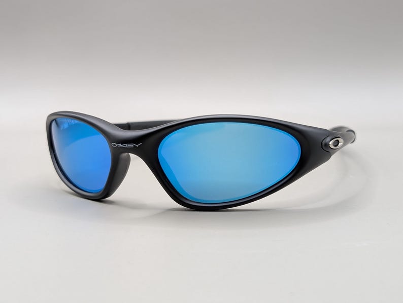 K&ouml;nnte beinhalten: Schwarze Sonnenbrille mit blau verspiegelten Gl&auml;sern. Die Sonnenbrille hat einen geschwungenen Rahmen und das Oakley-Logo ist auf der Seite sichtbar. Die Gl&auml;ser reflektieren das Licht und erzeugen ein hellblaues Aussehen.