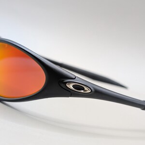 Pode incluir: &Oacute;culos de sol pretos com lentes laranja-avermelhadas. Os &oacute;culos de sol t&ecirc;m um design elegante e moderno com arma&ccedil;&atilde;o e hastes pretas. As lentes t&ecirc;m um efeito gradiente, passando do amarelo para o vermelho.