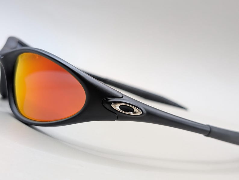 Pode incluir: &Oacute;culos de sol pretos com lentes laranja-avermelhadas. Os &oacute;culos de sol t&ecirc;m um design elegante e moderno com arma&ccedil;&atilde;o e hastes pretas. As lentes t&ecirc;m um efeito gradiente, passando do amarelo para o vermelho.
