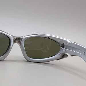 May include: Silver-framed sunglasses with dark lenses. The sunglasses have a sporty, wraparound design with a metallic finish. The temples are marked "Made in USA".