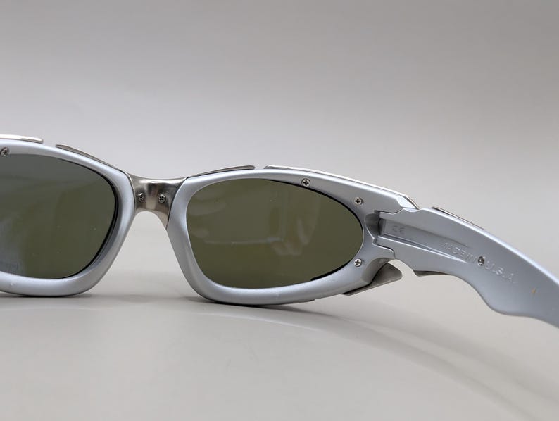 May include: Silver-framed sunglasses with dark lenses. The sunglasses have a sporty, wraparound design with a metallic finish. The temples are marked "Made in USA".