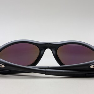 May include: Black sunglasses with purple-tinted lenses. The sunglasses are folded, showcasing the hinges and the sleek design. The lenses have a reflective quality, and the frame is matte black.