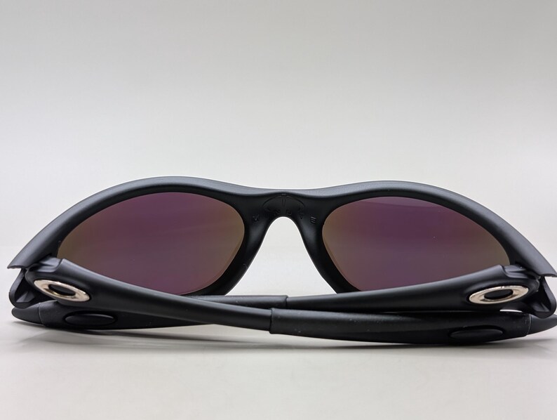 May include: Black sunglasses with purple-tinted lenses. The sunglasses are folded, showcasing the hinges and the sleek design. The lenses have a reflective quality, and the frame is matte black.