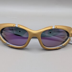 K&ouml;nnte beinhalten: Goldfarbene Sonnenbrille mit violetten Gl&auml;sern. Die Fassungen haben ein einzigartiges Design mit silbernen Akzenten und eine futuristische &Auml;sthetik. Die Sonnenbrille ist vor einem neutralen Hintergrund platziert.