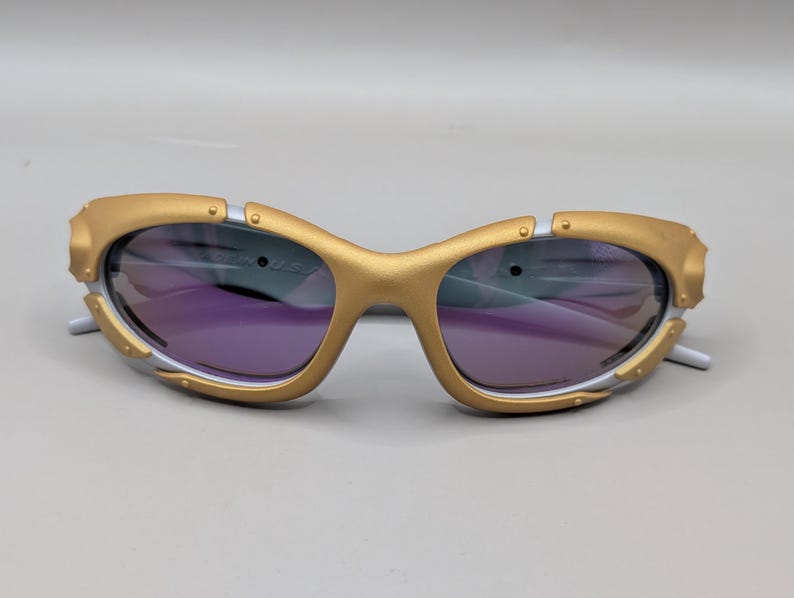 K&ouml;nnte beinhalten: Goldfarbene Sonnenbrille mit violetten Gl&auml;sern. Die Fassungen haben ein einzigartiges Design mit silbernen Akzenten und eine futuristische &Auml;sthetik. Die Sonnenbrille ist vor einem neutralen Hintergrund platziert.