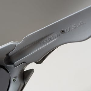 May include: Close-up of a pair of gray sunglasses. The text "MADE IN U.S.A." is printed on the arm of the sunglasses. The sunglasses have a sleek, modern design with a matte finish.