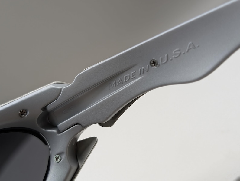 May include: Close-up of a pair of gray sunglasses. The text "MADE IN U.S.A." is printed on the arm of the sunglasses. The sunglasses have a sleek, modern design with a matte finish.
