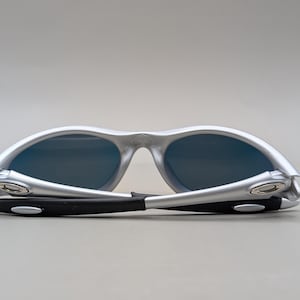 May include: Silver-framed sunglasses with dark blue lenses. The arms are black with silver accents. The sunglasses are folded, showcasing the design and style. A modern accessory for eye protection.
