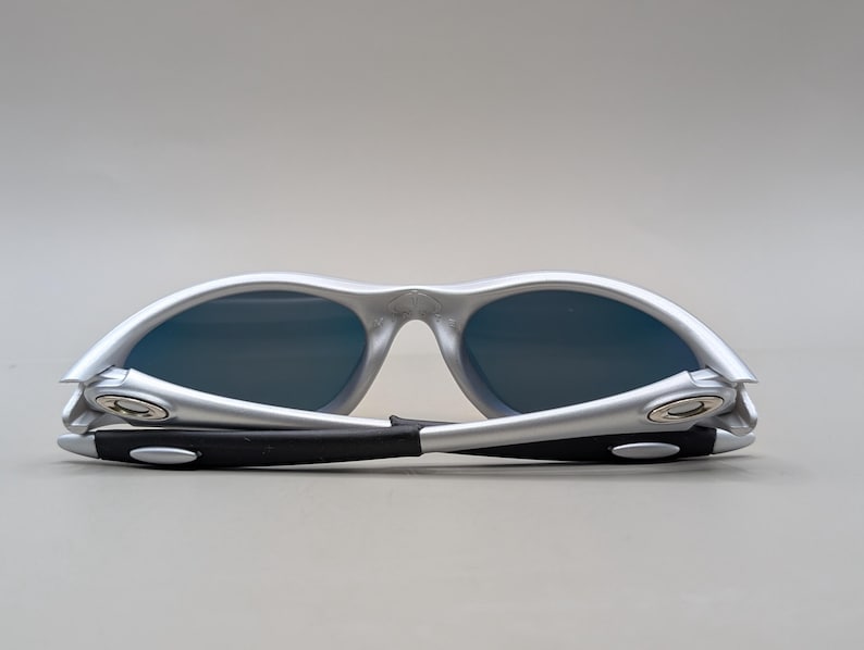May include: Silver-framed sunglasses with dark blue lenses. The arms are black with silver accents. The sunglasses are folded, showcasing the design and style. A modern accessory for eye protection.