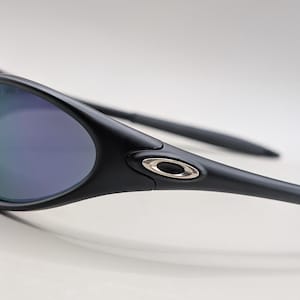May include: Close-up of black sunglasses with a curved frame and reflective lenses. The lenses have a gradient of green and purple hues. The temples are sleek and feature a silver accent near the hinge.