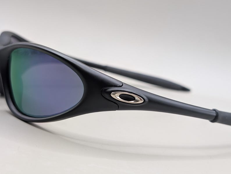 May include: Close-up of black sunglasses with a curved frame and reflective lenses. The lenses have a gradient of green and purple hues. The temples are sleek and feature a silver accent near the hinge.