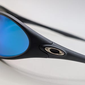 May include: Close-up of black sunglasses with blue mirrored lenses. The sunglasses have a sleek, modern design with a silver accent on the temple. The lenses reflect a vibrant blue color, contrasting with the black frame.