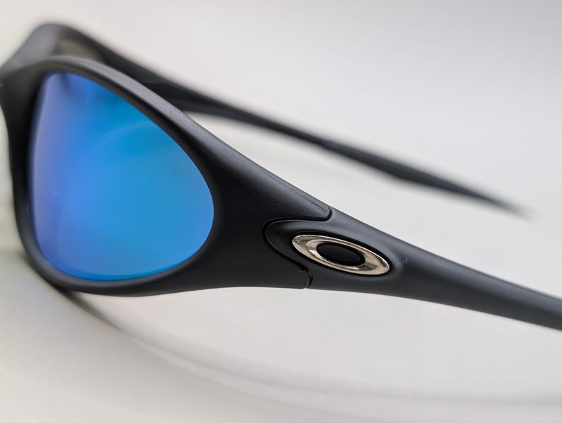 May include: Close-up of black sunglasses with blue mirrored lenses. The sunglasses have a sleek, modern design with a silver accent on the temple. The lenses reflect a vibrant blue color, contrasting with the black frame.