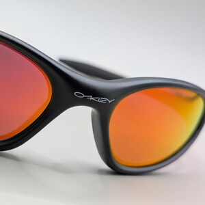Pode incluir: Close-up de &oacute;culos de sol pretos com lentes vibrantes laranja e vermelhas. Os &oacute;culos de sol t&ecirc;m um design elegante e moderno com o nome da marca "Oakley" vis&iacute;vel na arma&ccedil;&atilde;o. As lentes fazem uma transi&ccedil;&atilde;o do vermelho para o laranja.