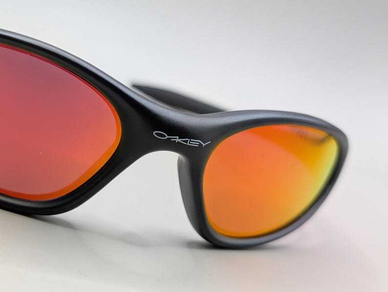Pode incluir: Close-up de &oacute;culos de sol pretos com lentes vibrantes laranja e vermelhas. Os &oacute;culos de sol t&ecirc;m um design elegante e moderno com o nome da marca "Oakley" vis&iacute;vel na arma&ccedil;&atilde;o. As lentes fazem uma transi&ccedil;&atilde;o do vermelho para o laranja.
