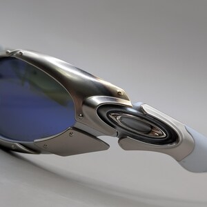 May include: Close-up of silver sunglasses with blue lenses. The sunglasses have a futuristic design with a metallic frame and white arms. The lenses are a vibrant blue colour.