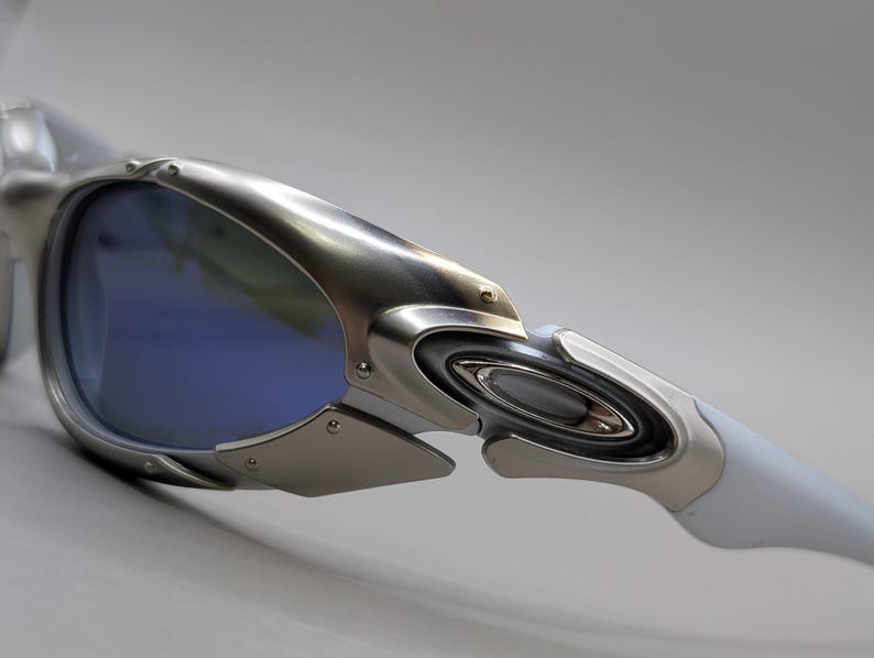 May include: Close-up of silver sunglasses with blue lenses. The sunglasses have a futuristic design with a metallic frame and white arms. The lenses are a vibrant blue colour.