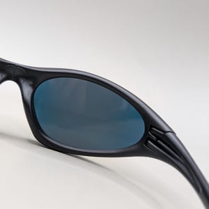 Pode incluir: Close-up de &oacute;culos de sol pretos com lentes azul escuro. Os &oacute;culos de sol t&ecirc;m um design elegante e curvo com um acabamento fosco. As hastes dos &oacute;culos de sol s&atilde;o ligeiramente curvas e apresentam um detalhe canelado perto das dobradi&ccedil;as. As lentes s&atilde;o ovais.