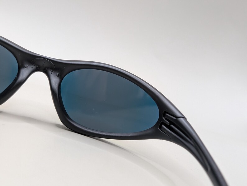 Pode incluir: Close-up de &oacute;culos de sol pretos com lentes azul escuro. Os &oacute;culos de sol t&ecirc;m um design elegante e curvo com um acabamento fosco. As hastes dos &oacute;culos de sol s&atilde;o ligeiramente curvas e apresentam um detalhe canelado perto das dobradi&ccedil;as. As lentes s&atilde;o ovais.
