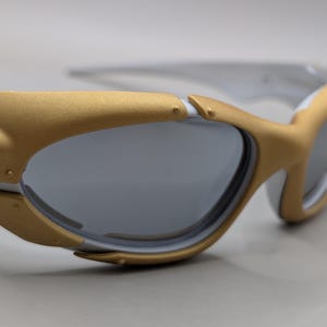May include: White and gold-framed sunglasses with gray lenses. The sunglasses have a sporty design with a gold accent on the frame and temples. The lenses are a dark gray color, providing protection from the sun.