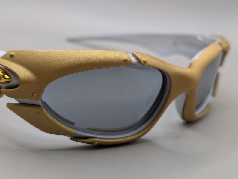 May include: White and gold-framed sunglasses with gray lenses. The sunglasses have a sporty design with a gold accent on the frame and temples. The lenses are a dark gray color, providing protection from the sun.