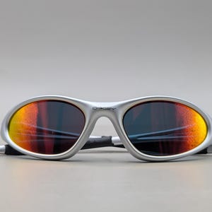 May include: Silver-framed sunglasses with orange and yellow mirrored lenses. The sunglasses have a sleek, sporty design with the brand name "Oakley" visible on the frame. The lenses are oval-shaped.