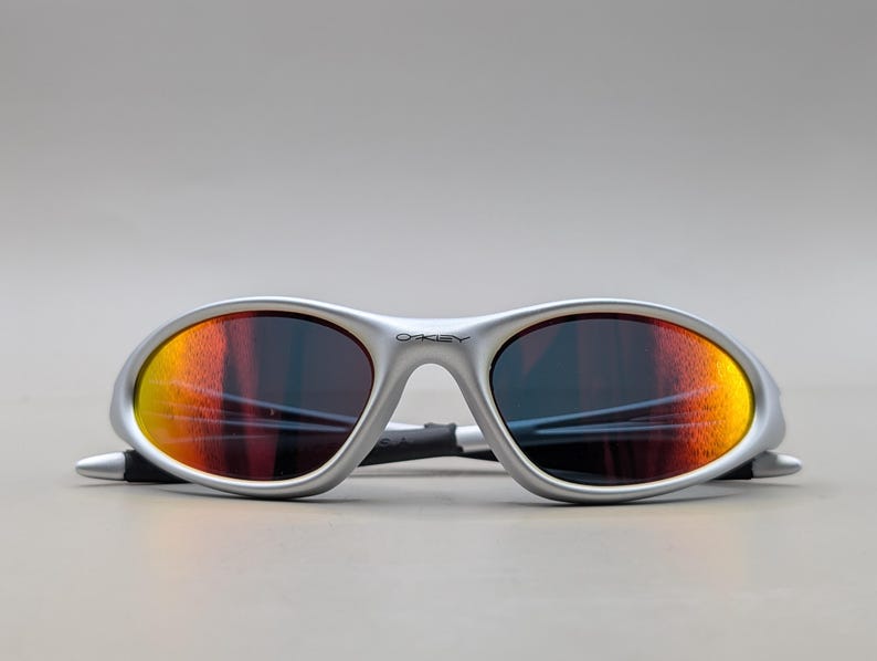 May include: Silver-framed sunglasses with orange and yellow mirrored lenses. The sunglasses have a sleek, sporty design with the brand name "Oakley" visible on the frame. The lenses are oval-shaped.