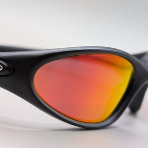 Pode incluir: Close-up de &oacute;culos de sol pretos com lentes gradientes laranja e amarelo vibrantes. Os &oacute;culos de sol t&ecirc;m um design elegante e moderno com um acabamento fosco. As lentes s&atilde;o curvas, oferecendo um amplo campo de vis&atilde;o.