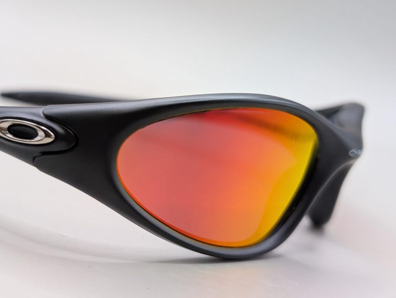 Pode incluir: Close-up de &oacute;culos de sol pretos com lentes gradientes laranja e amarelo vibrantes. Os &oacute;culos de sol t&ecirc;m um design elegante e moderno com um acabamento fosco. As lentes s&atilde;o curvas, oferecendo um amplo campo de vis&atilde;o.